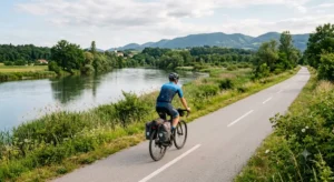 Kolesar na urejeni ravninski kolesarski poti ob reki Savi v okolici Dobove s pogledom na zeleno naravo in hribe v ozadju.